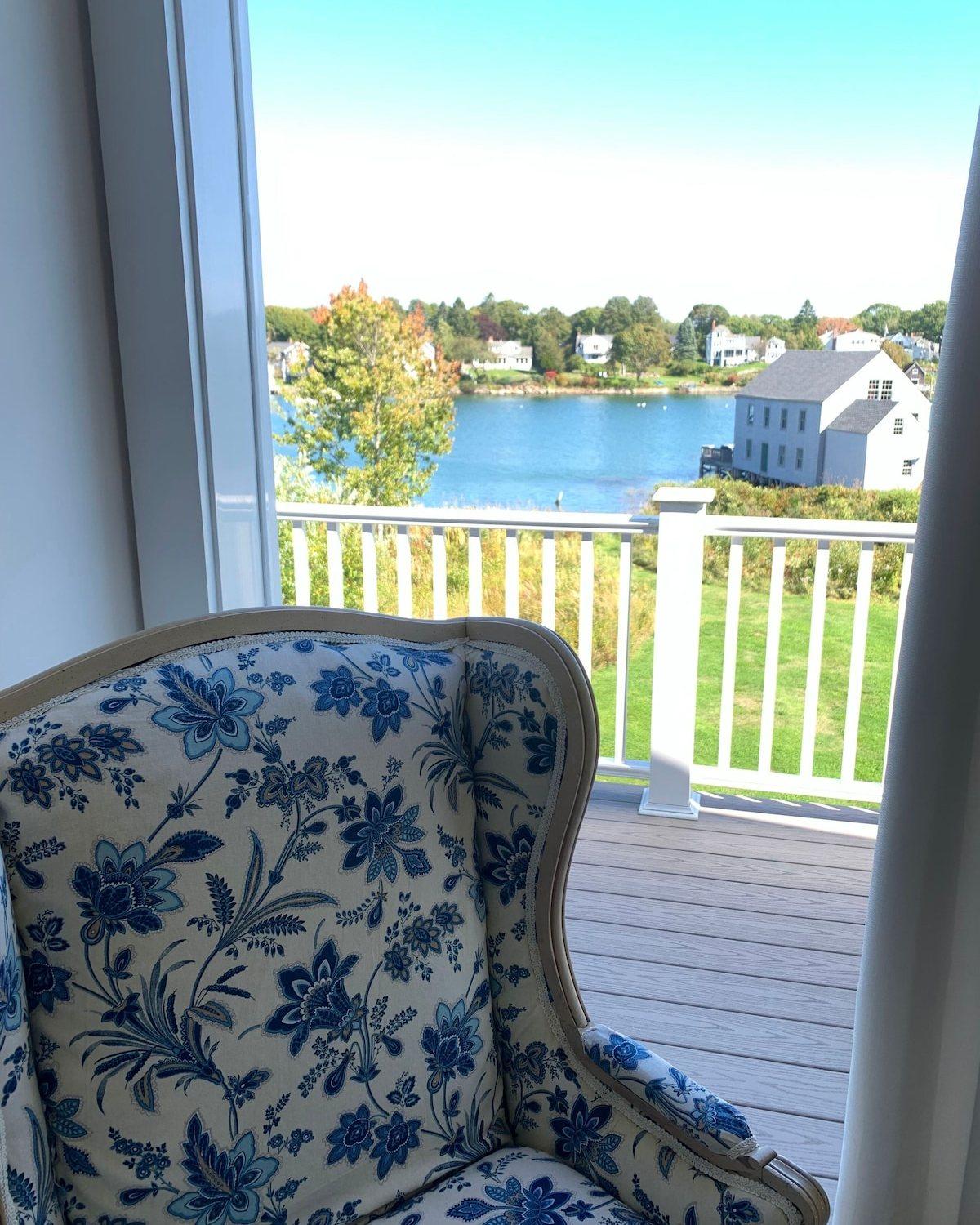 Blue-and-white wing chair beside an open door onto the upstairs balcony, with its white railing, a white cottage, and the harbor beyond.