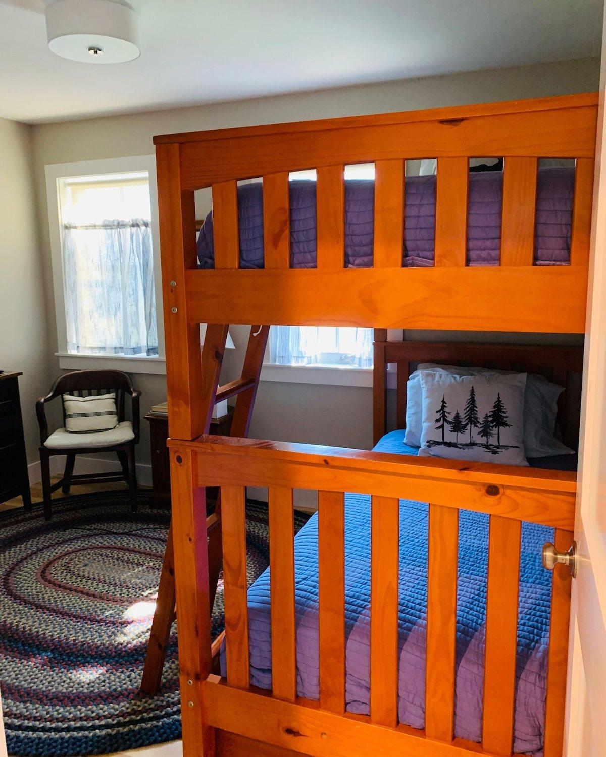 Bedroom with a wooden bunk bed, blue quilts, a braided rug, and a chair by the window.