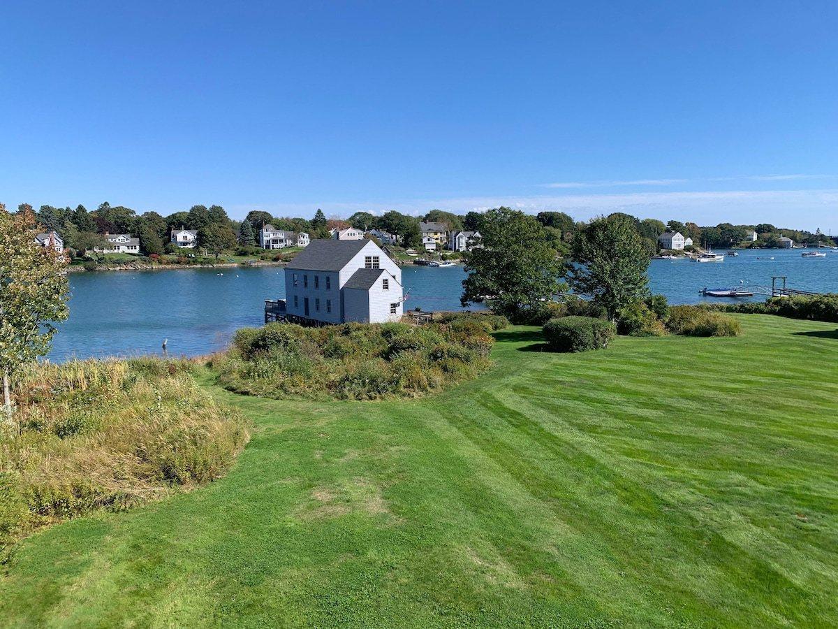Mown lawn running down to the cove, with a white fish house on pilings and boats moored beyond.