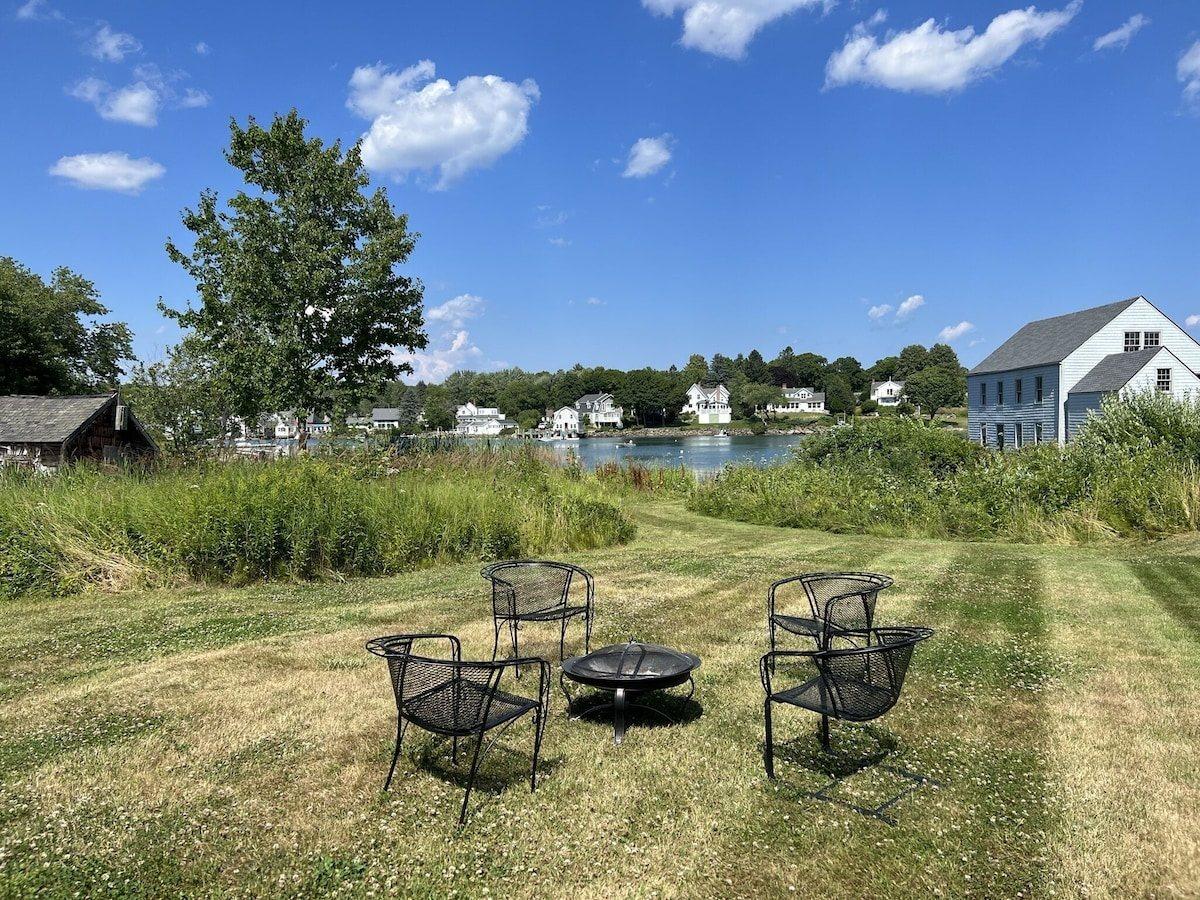 Fire pit and four metal chairs on the lawn, with the harbor and village houses beyond the marsh grass.