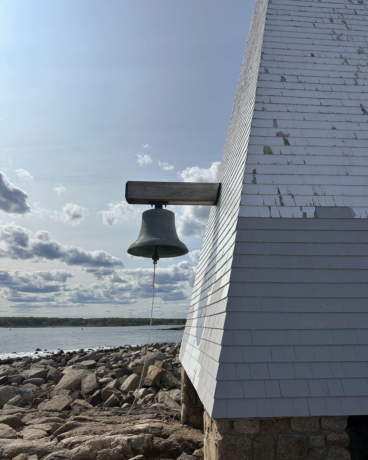The bronze fog bell hanging from a wooden arm on the white-shingled wall of Goat Island Light, with granite rocks and the harbor behind it.