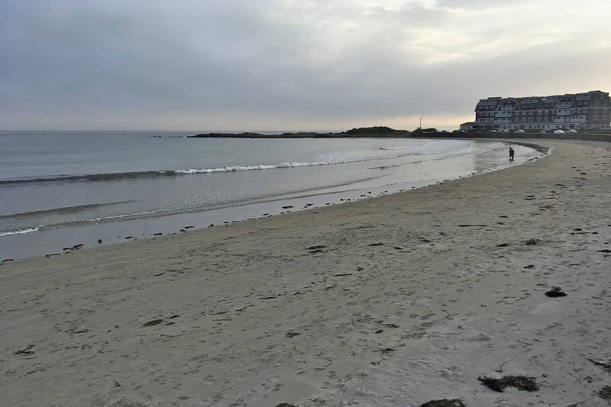Gooch's Beach in Kennebunk on an overcast day: wet sand, small waves, and a rocky point with buildings in the distance.