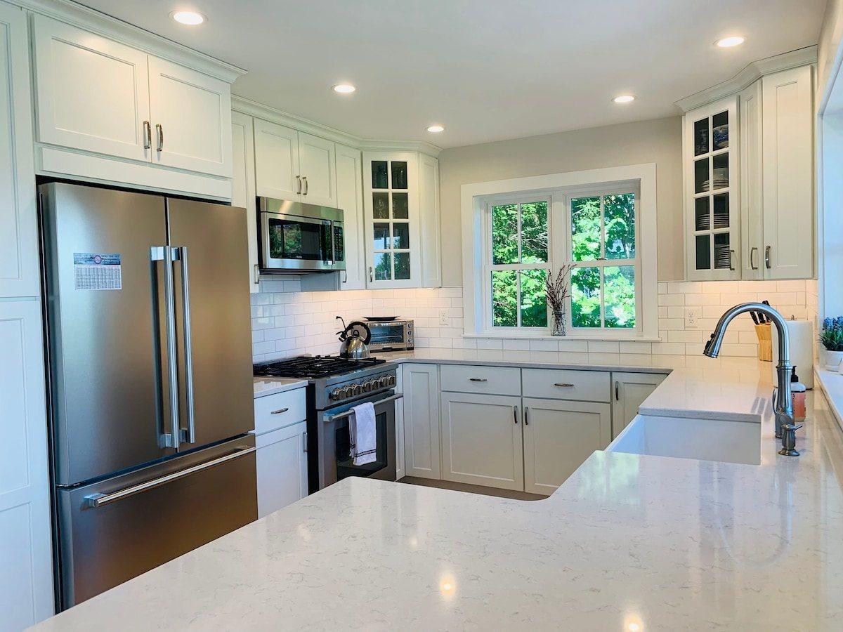 White kitchen with a gas range, large refrigerator, farmhouse sink under the window, and a long stone counter.