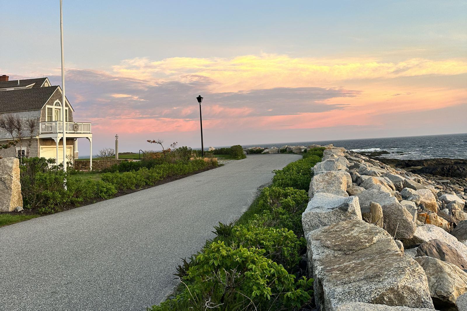 A paved path along the seawall at South Point, Biddeford Pool, with a shingled house on the left and the ocean under a pink evening sky.